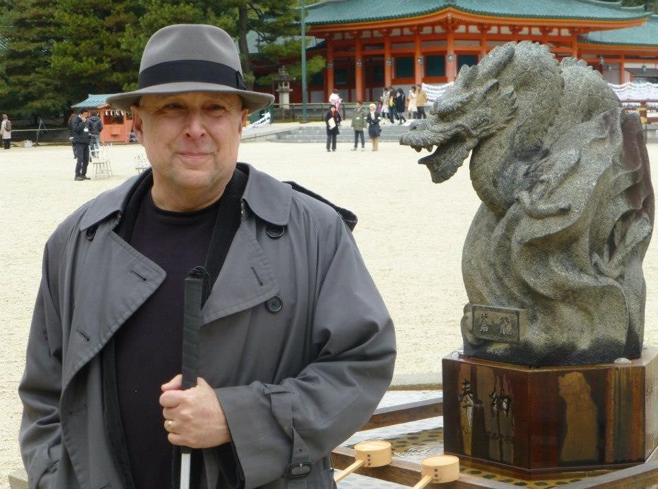 Stephen Kuusisto wearing a fedora and trench coat, standing beside a dragon in Kyoto, Japan